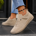 Beige orthopedic sneakers for women with elastic strap and stitched sole, worn with frayed jeans on dark tiles.