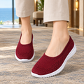 Red knitted slip-on sneakers with a white sole, worn with beige trousers on a carpet by a window.
