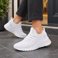 Knit sneakers women can wear daily: a white lace-up pair in breathable knitted mesh, worn with black trousers on a grey pavement.