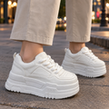 White platform sneakers with thick braided laces and a chunky sole, worn with beige trousers on a pavement at sunset