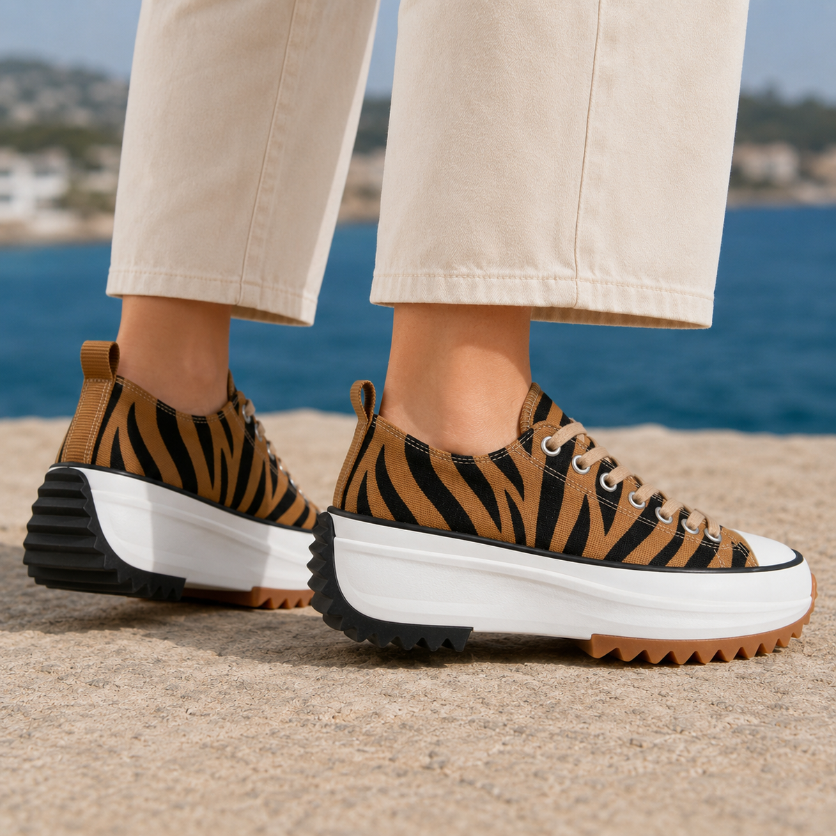 Orthopedic sneakers for women in brown and black tiger-stripe woven style with a white chunky sole, worn with cream trousers on a sandy ledge