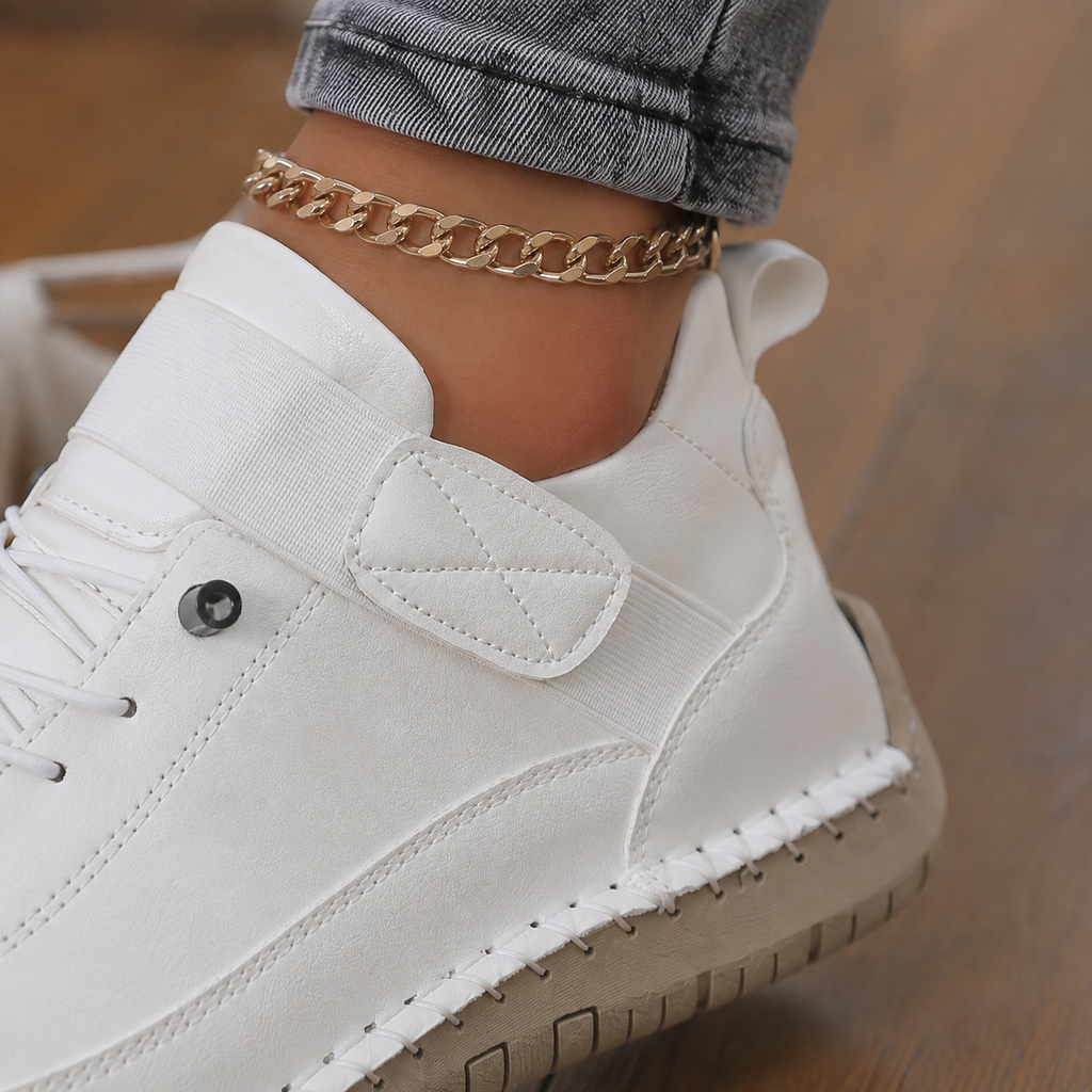 Close-up of a white leather sneaker showing the elastic strap with stitched tab, eyelet, seams and beige sole