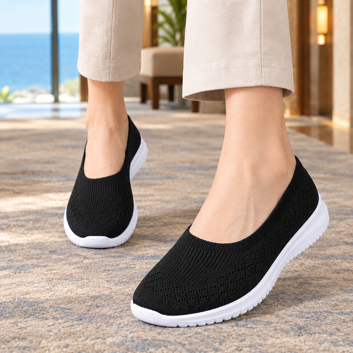 Black knitted slip on trainers with a white sole, worn with beige trousers on a rug.