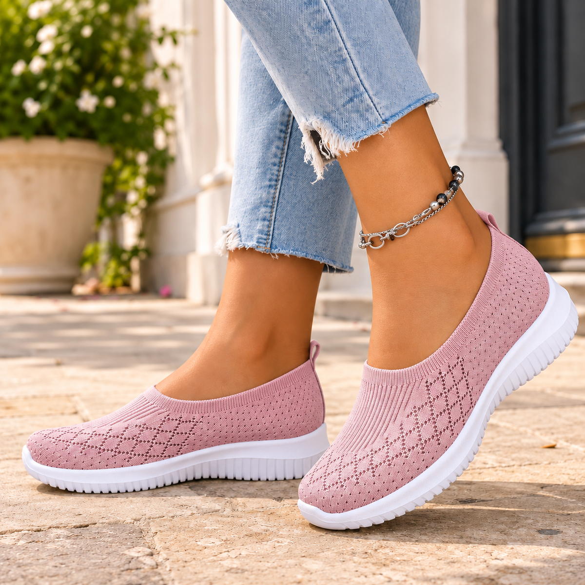 Pink knitted slip on trainers with a white sole, worn with jeans on a stone pavement.