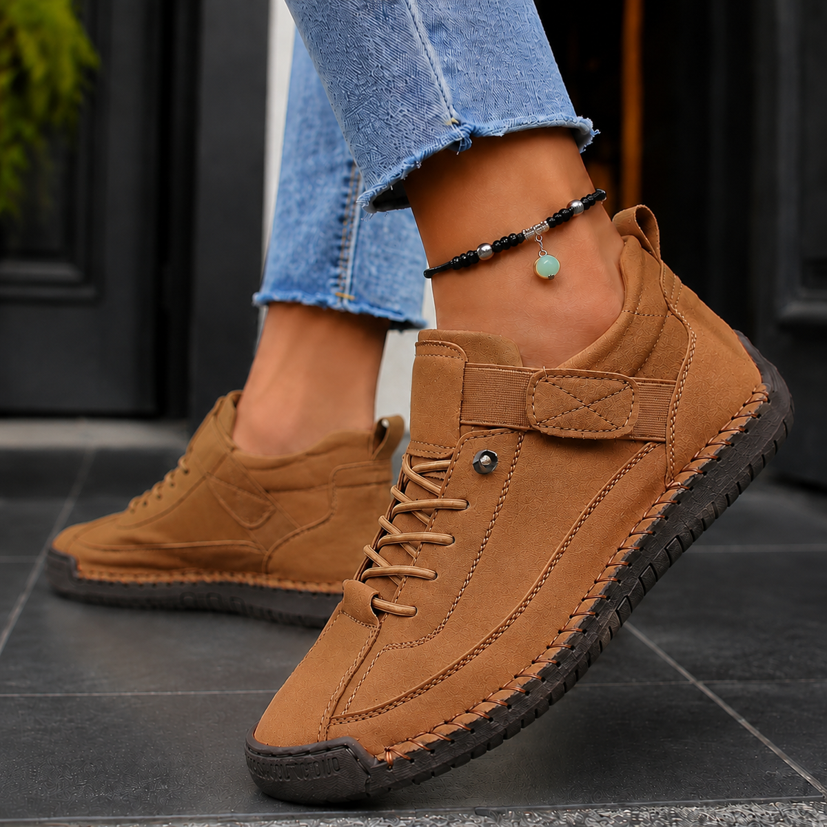 Khaki-brown lace-up sneakers with elastic strap and dark stitched sole, worn with jeans.