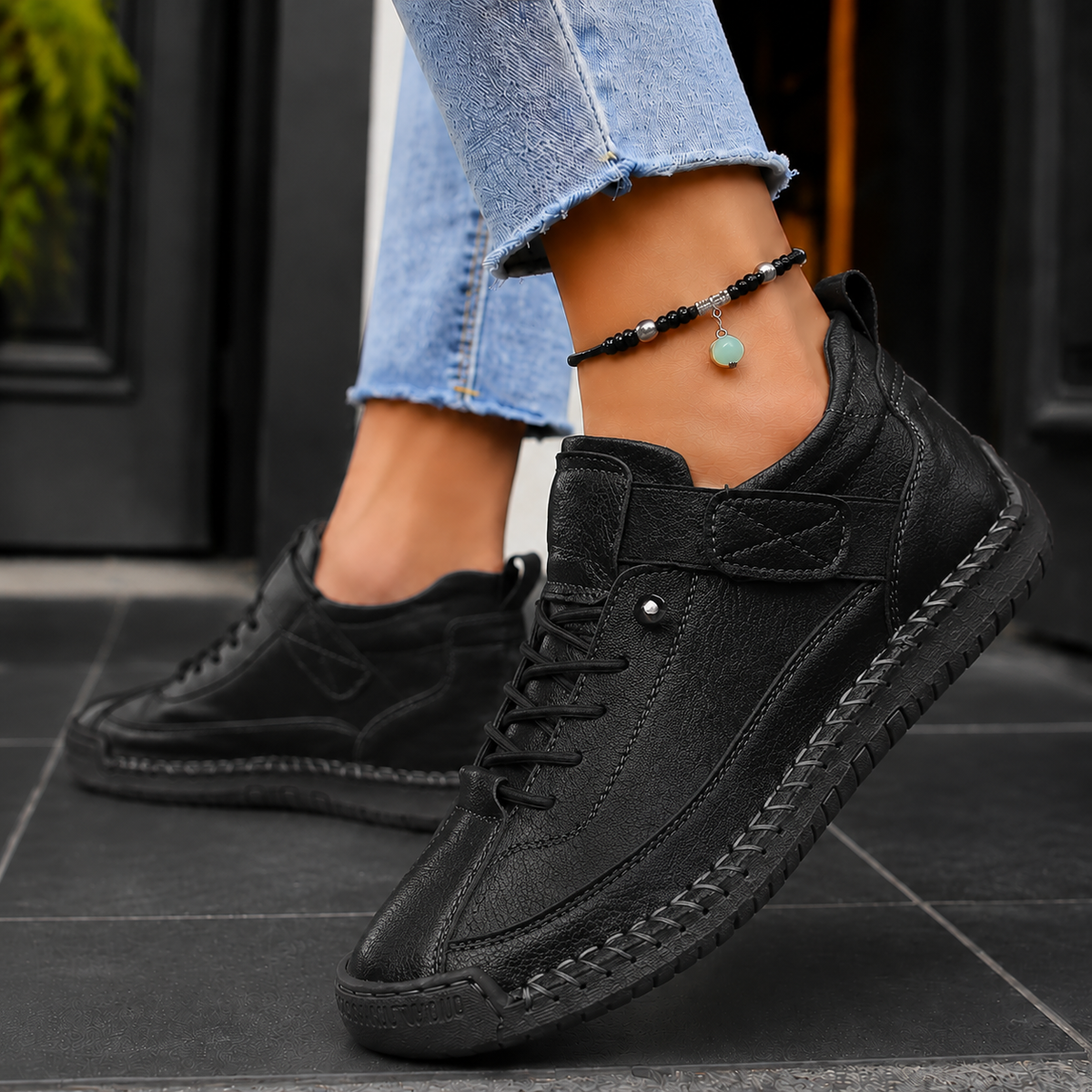 Black lace-up sneakers with strap and matching black stitched sole, worn with frayed jeans.
