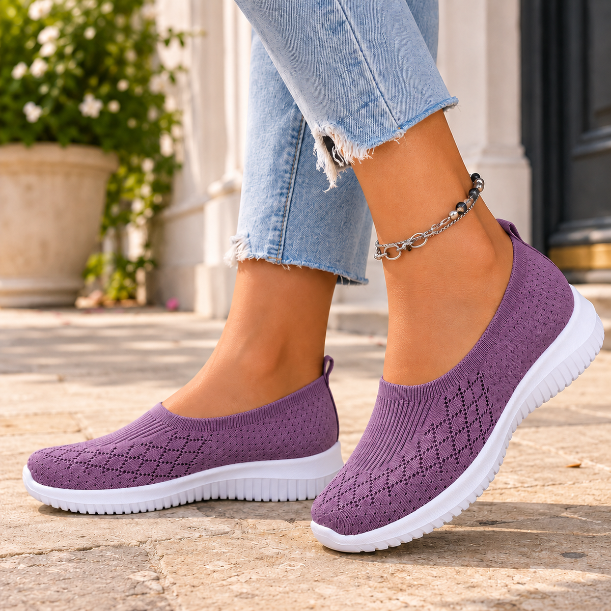 Purple knitted slip on trainers with a white sole, worn with jeans on a stone pavement.