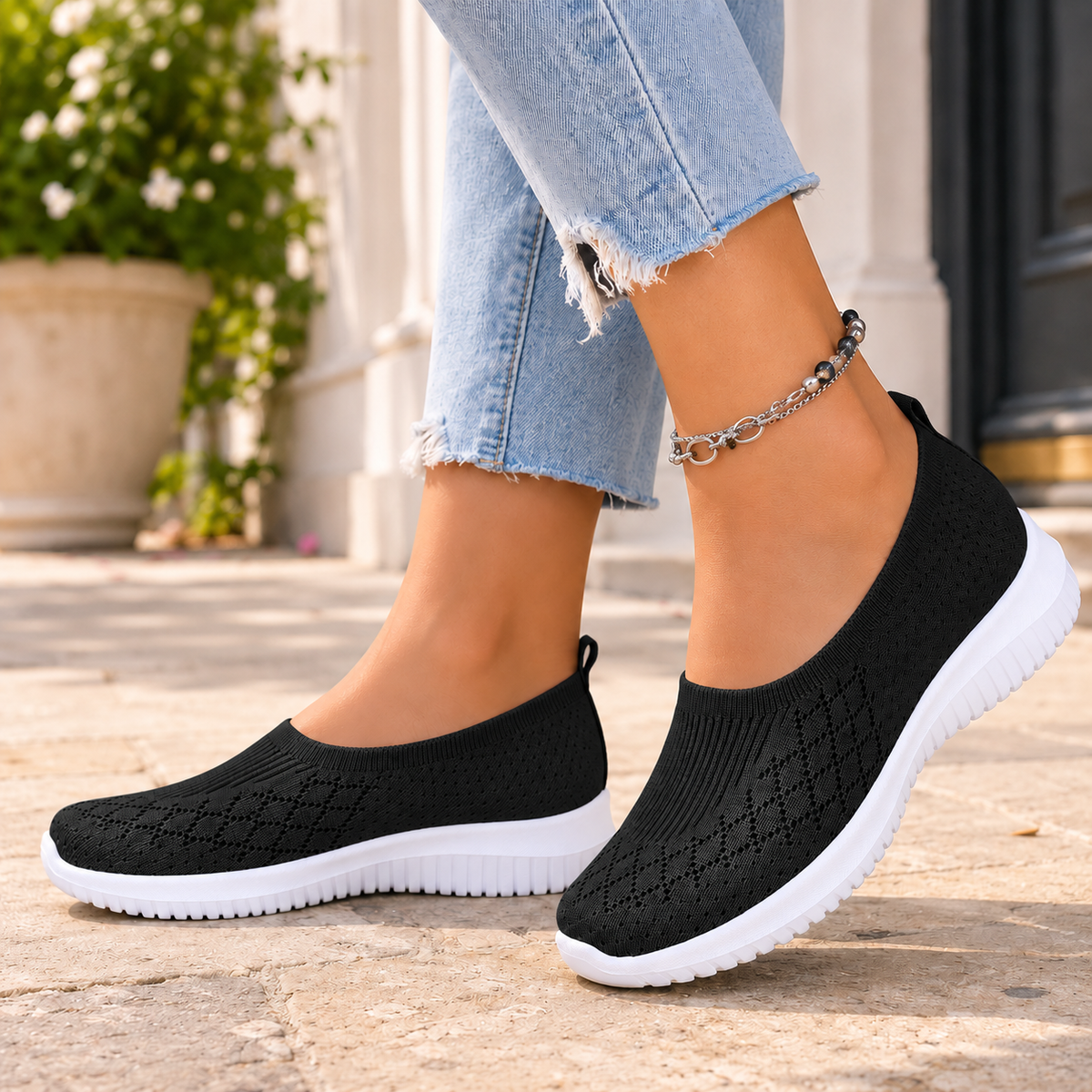 Black knitted slip on trainers with a white sole, worn with jeans on a stone pavement.