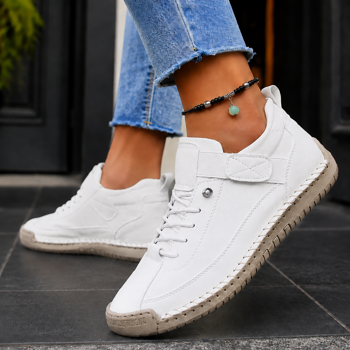 White lace-up sneakers with elastic strap and light grey stitched sole, worn with jeans on dark tiles.